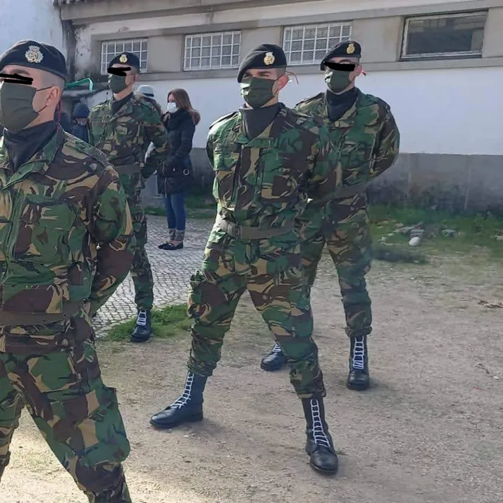 Four soldiers in camouflage and berets, marching in formation across a barracks yard in cloth face masks.