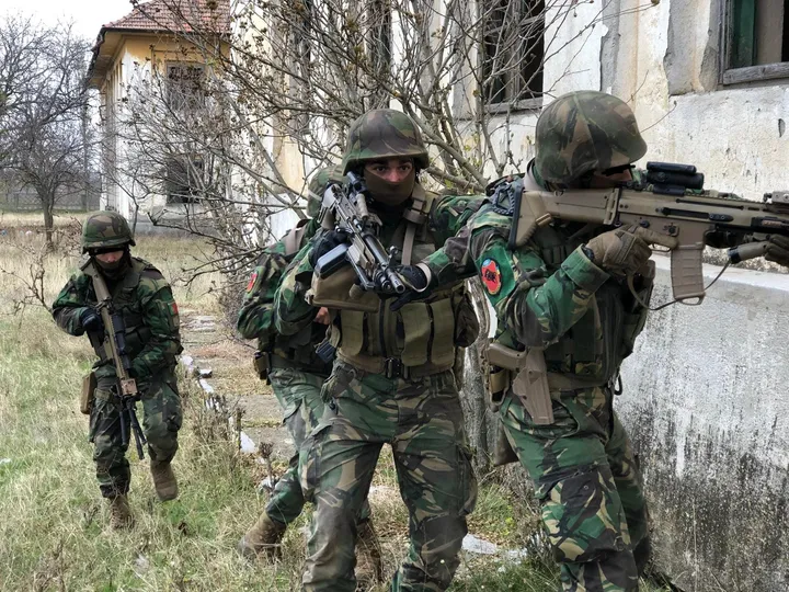 Four soldiers in helmets and Portuguese camouflage stacked along the wall of a derelict building on an exercise, rifles up.