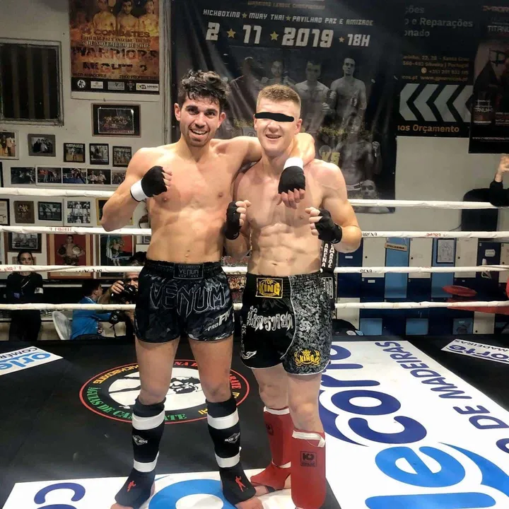 Two fighters in shorts and shin pads standing arm in arm in the ring after their bout, hands still wrapped, under a banner for a November 2019 kickboxing card.